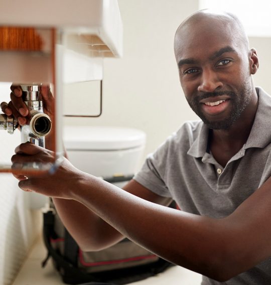 Young black male plumber sitting on the floor fixing a bathroom sink, looking to camera, close up